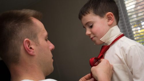 A young dad helps his son get ready by helping him tie his neck tie.